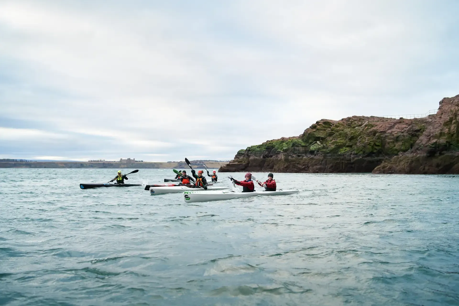 Kayakers paddling across a choppy lake on an overcast day, near a rocky shoreline.