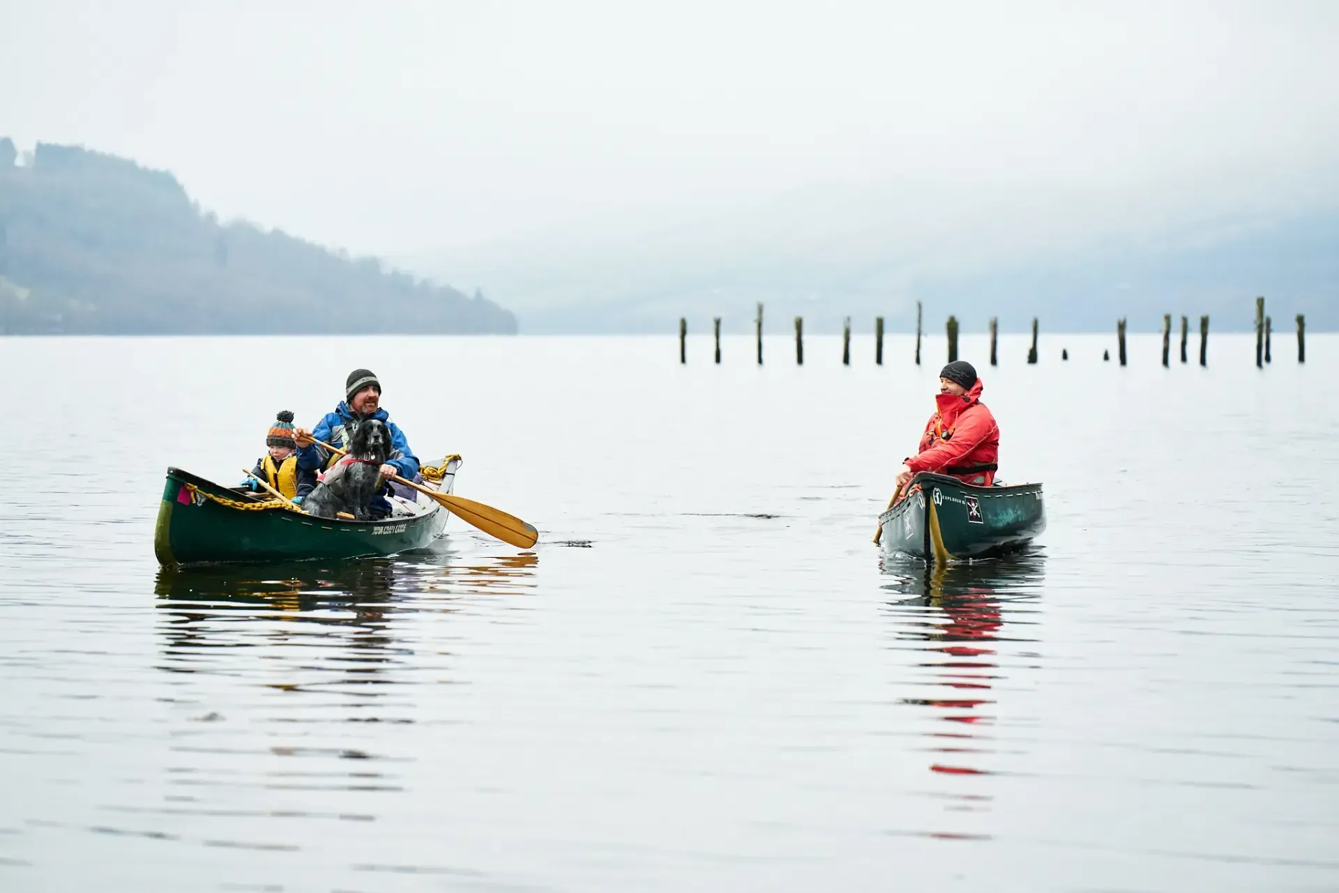 Two canoes on a calm lake. One holds two people paddling, the other has one person sitting. Gray sky and distant shore.