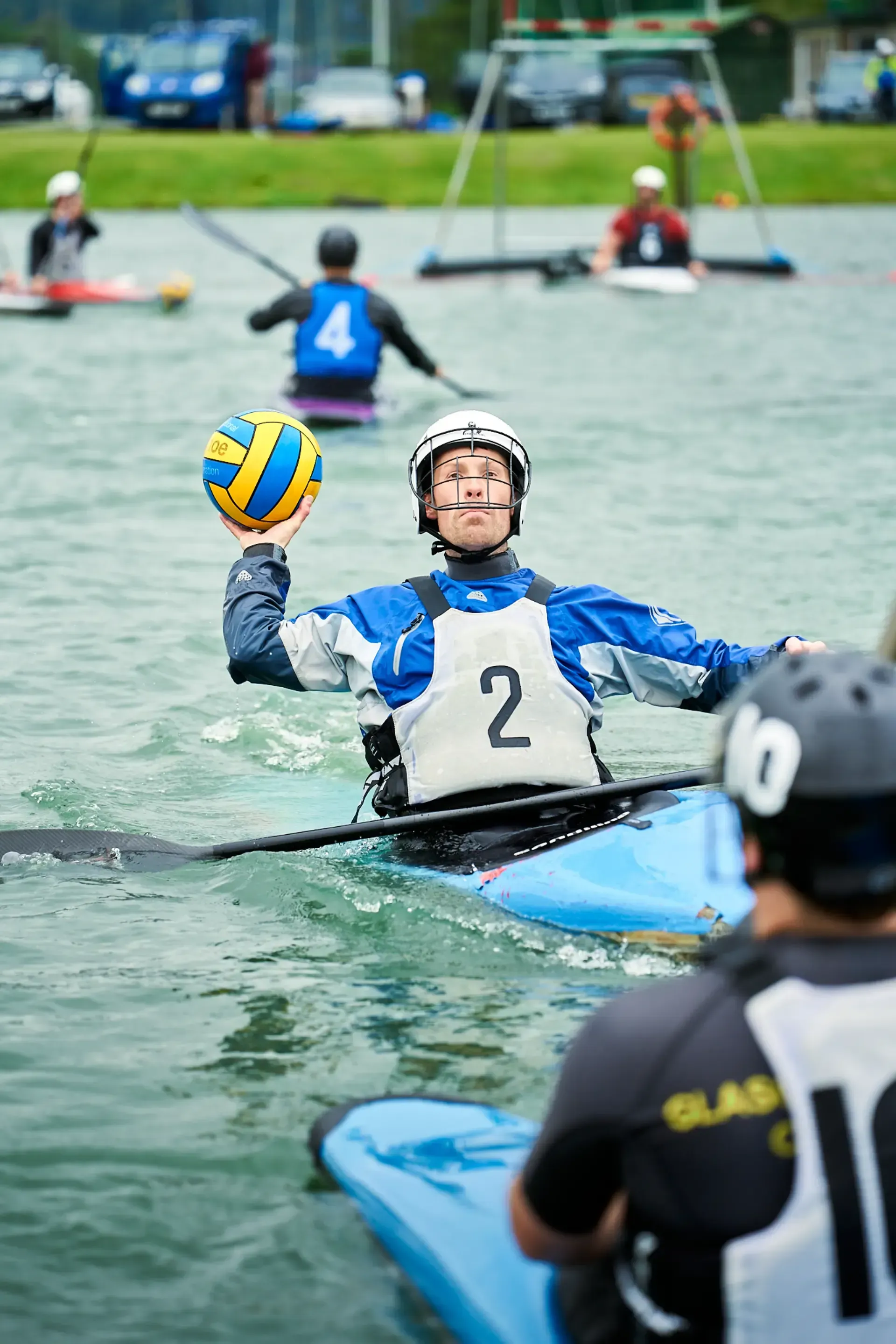 A kayaker in a blue and white jersey with the number 2 prepares to throw a yellow and blue ball. Other kayakers are in the water nearby.