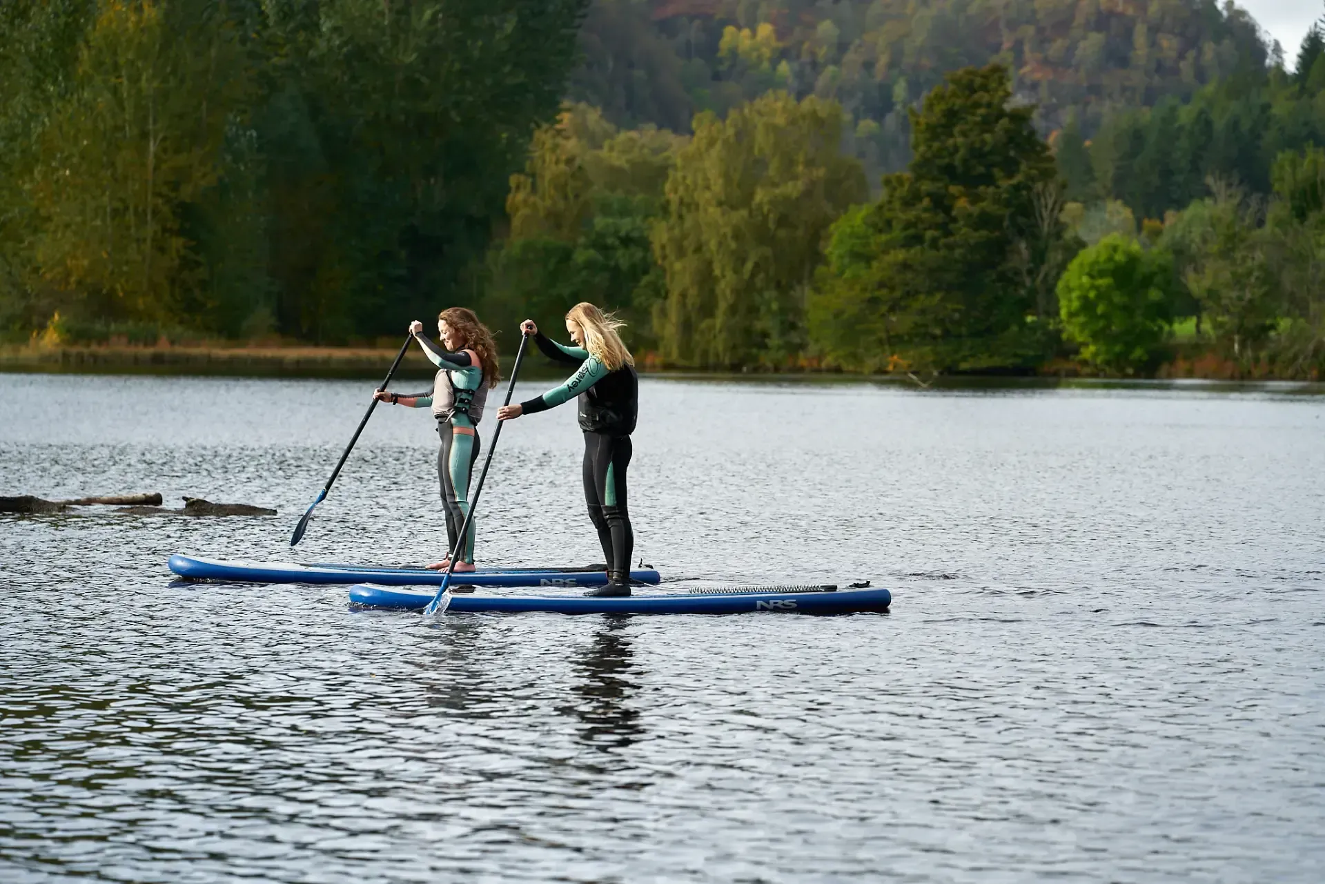 Two women on paddleboards on a lake, paddling. They are wearing wetsuits, trees and overcast sky in the background.