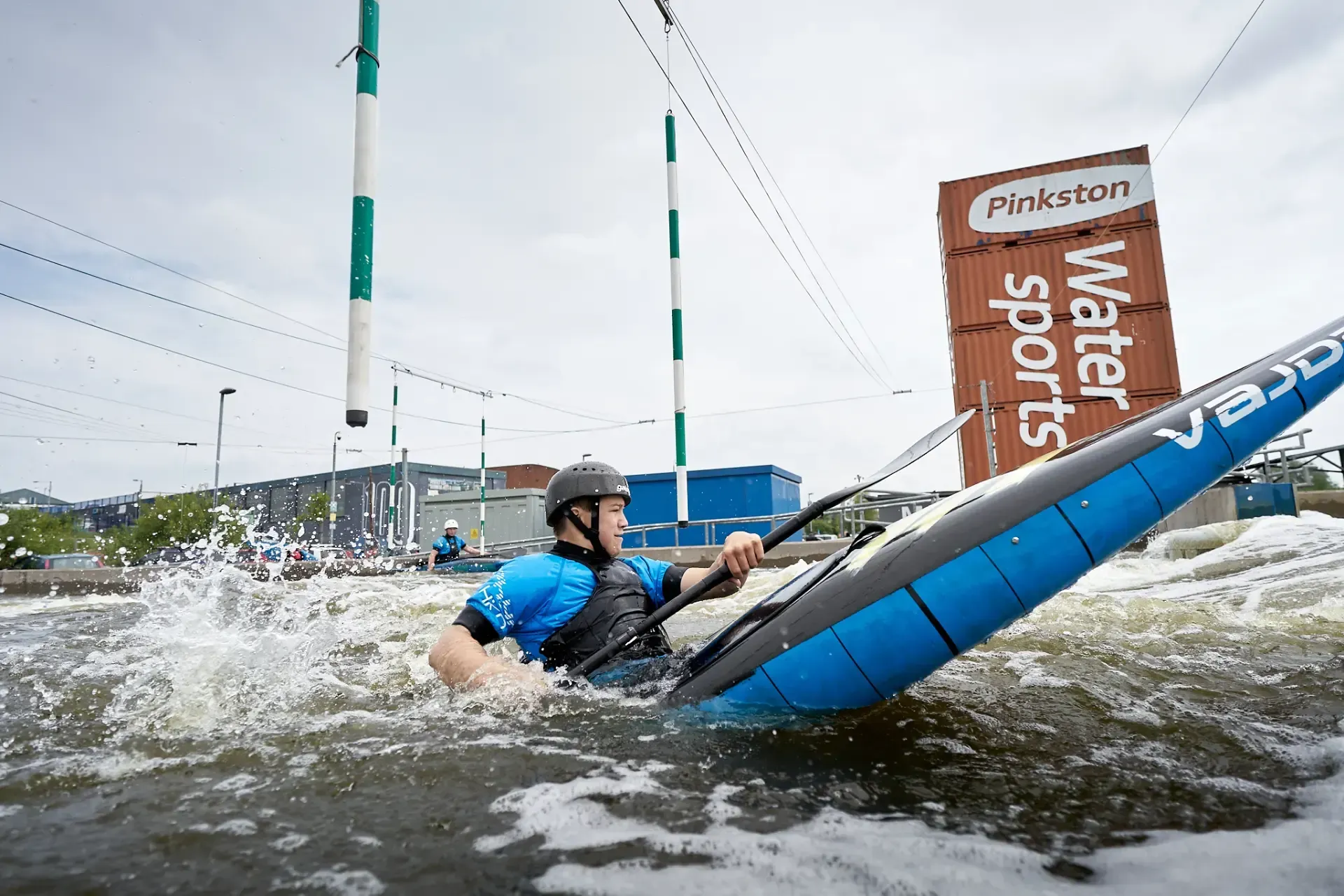 A kayaker navigates whitewater rapids, heading toward a gate at Pinkston Watersports. He wears a helmet and blue top.