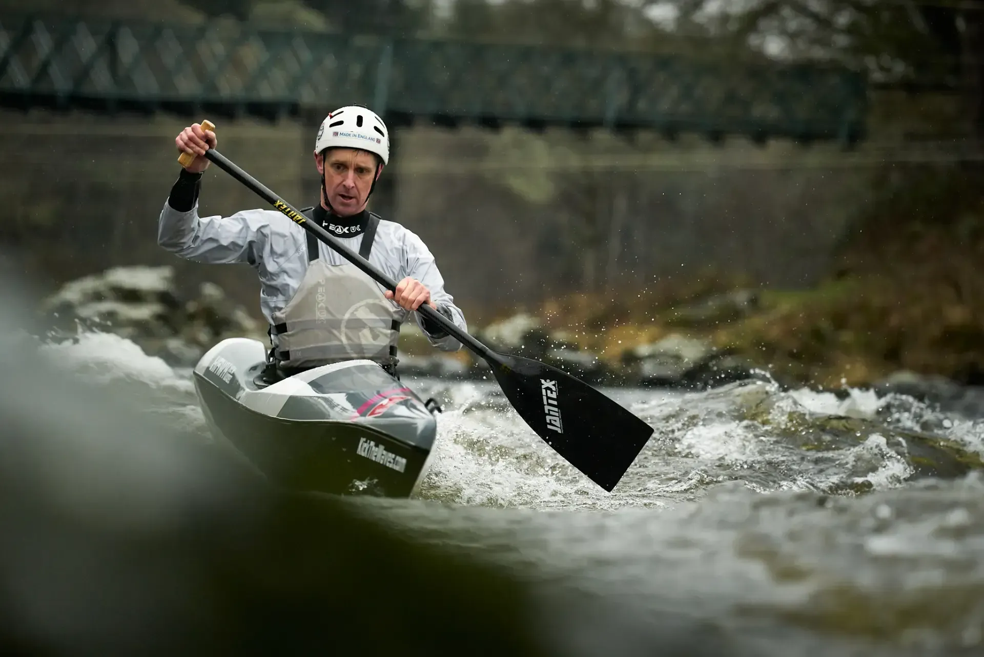 Man in a kayak paddles on a river, wearing a helmet, near a bridge.