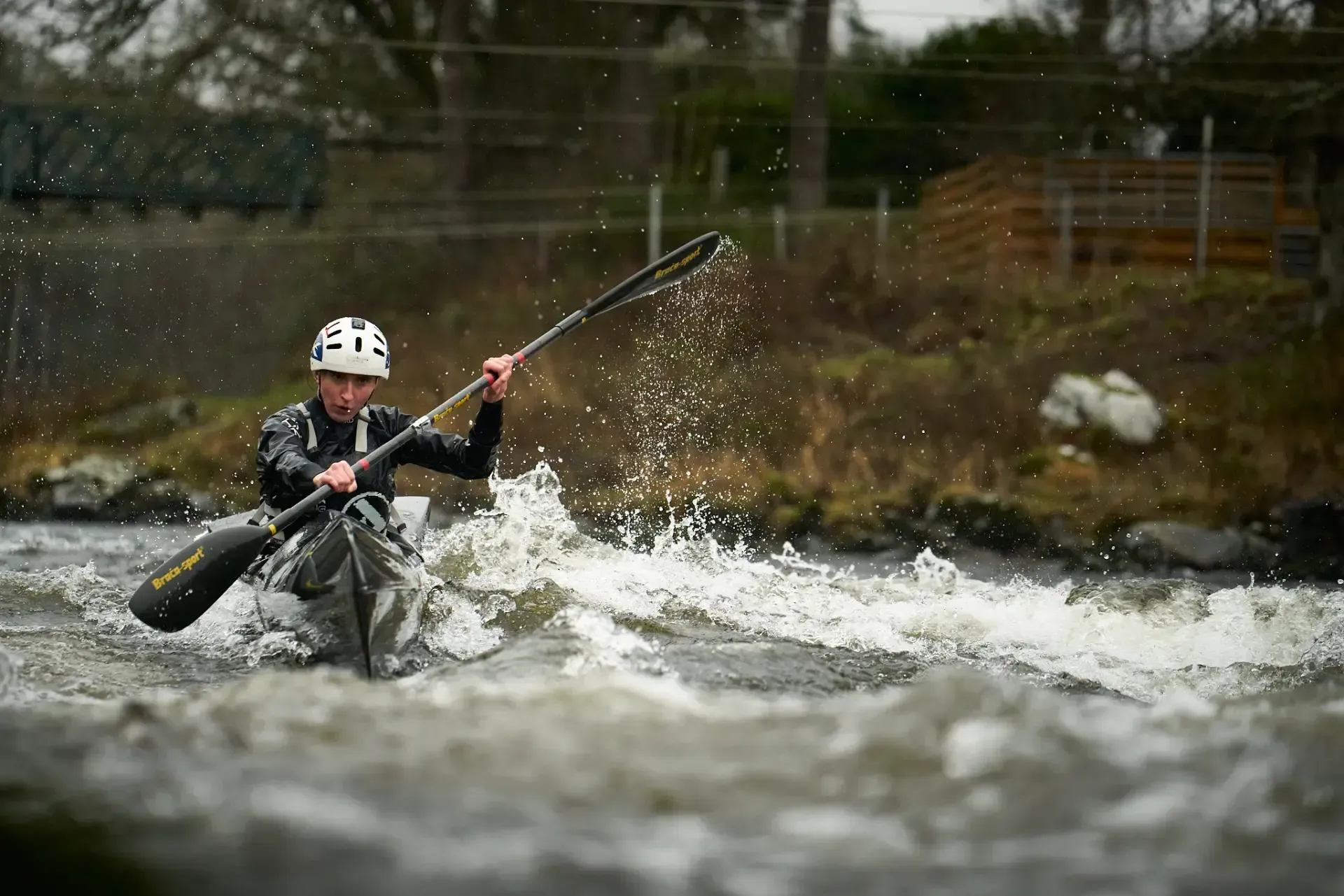A kayaker navigates white water rapids, paddling with a two-bladed oar. They wear a helmet and dark clothing.