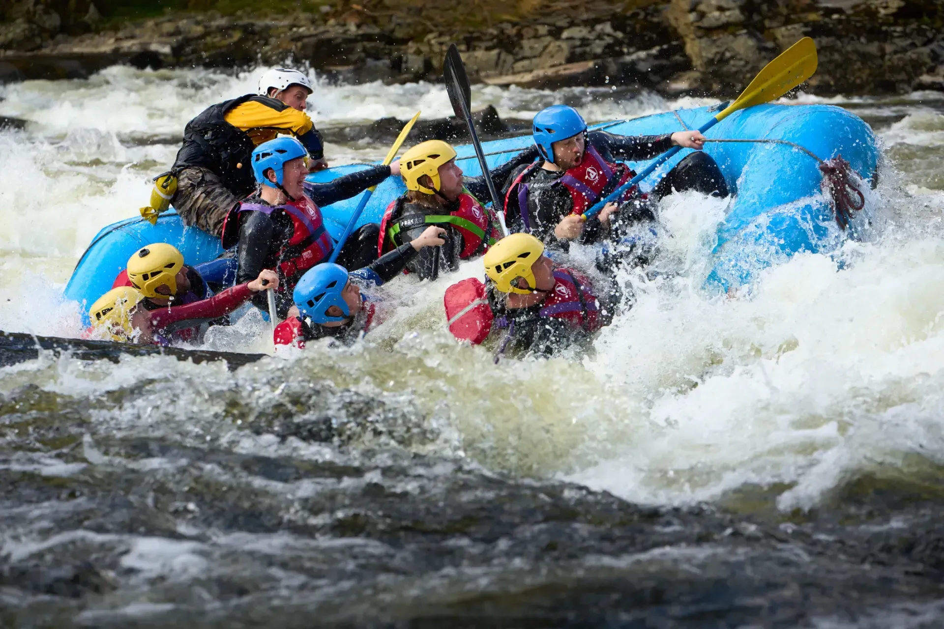 Rafting through whitewater rapids. A blue raft carries eight people in helmets and life vests, paddling aggressively through churning water.