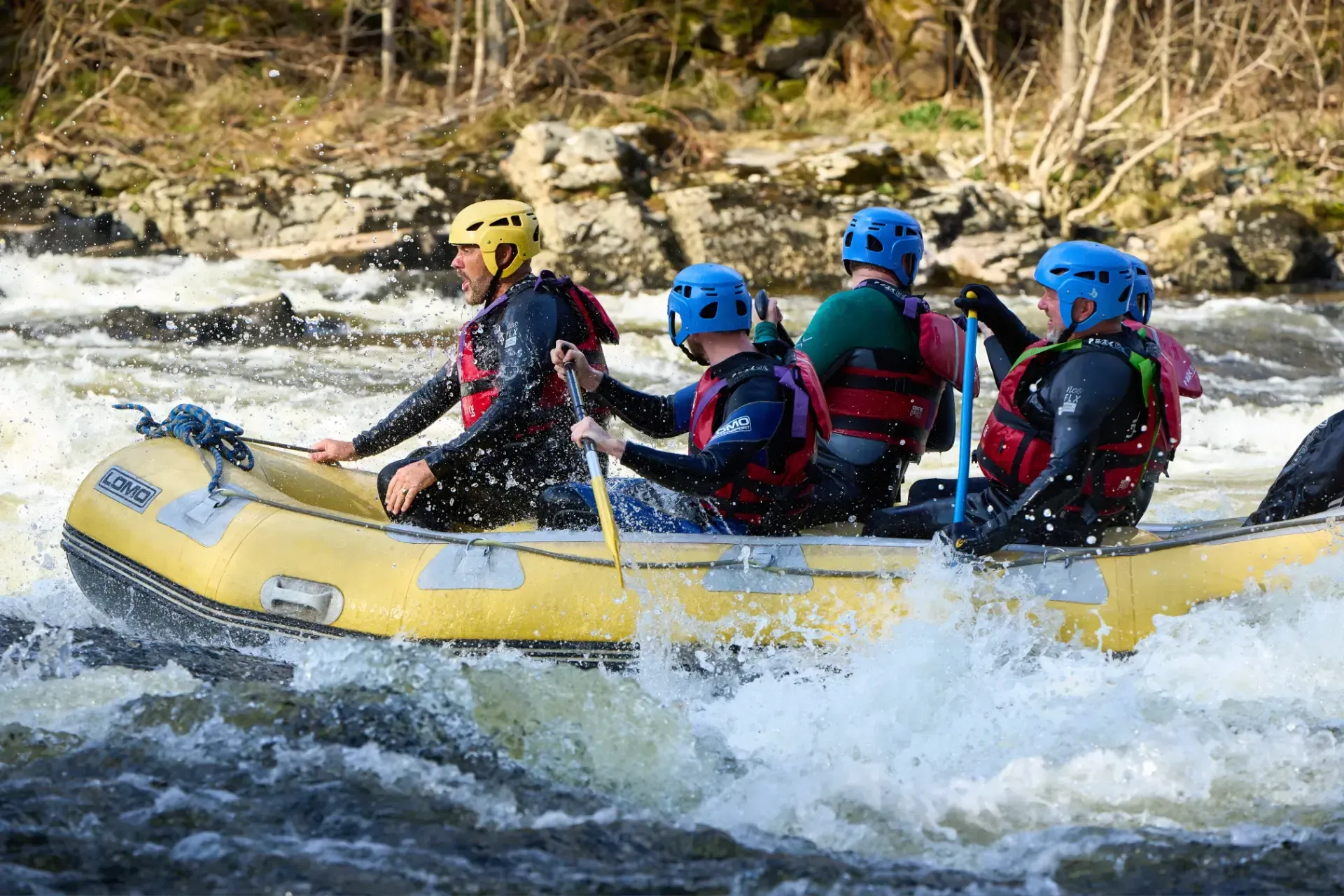 A group of four people in a yellow raft navigate whitewater rapids. They wear helmets, life vests, and wetsuits, paddling furiously.