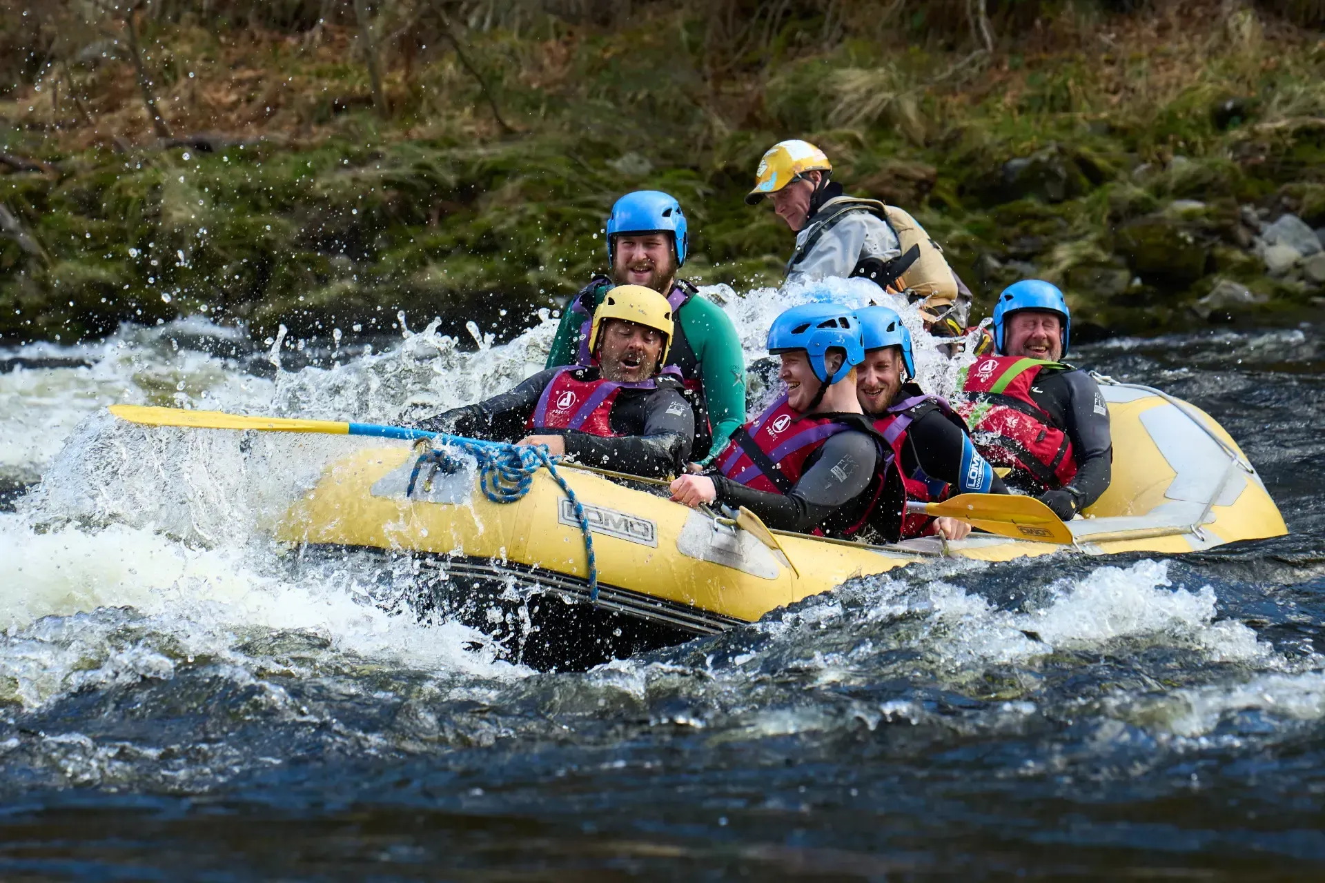 People rafting on a yellow inflatable boat through whitewater rapids. They wear helmets and life vests, splashing water.