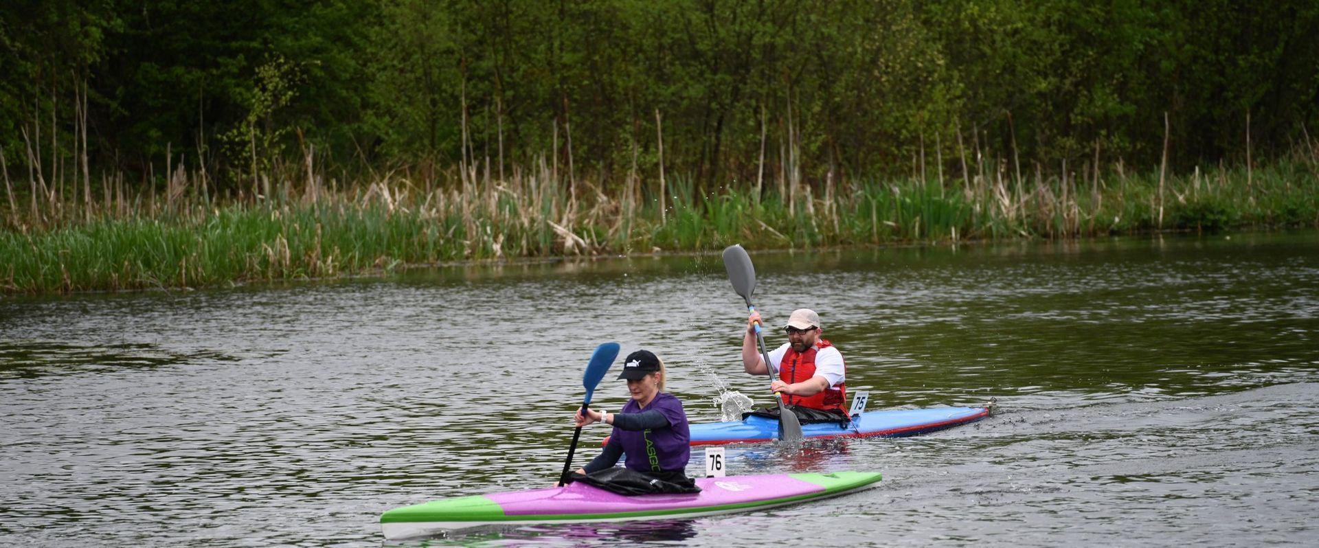 Two people kayaking on a lake with green and pink kayaks, surrounded by lily pads and trees.