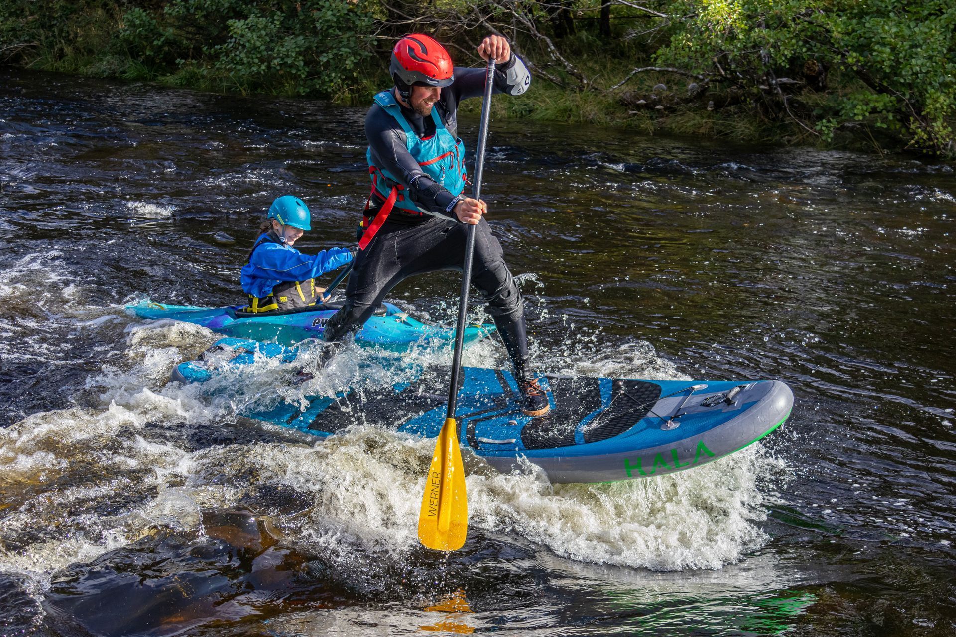 Person stand-up paddleboarding on a whitewater river, another person in a kayak behind them. Both wear safety gear, red and blue helmets.