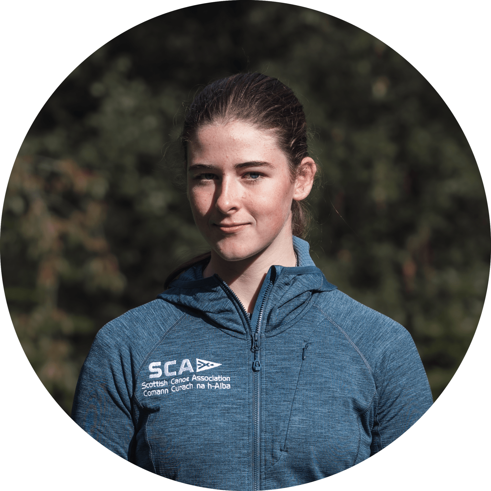 A young woman in a blue hoodie with a SCA logo is smiling in a natural outdoor setting.
