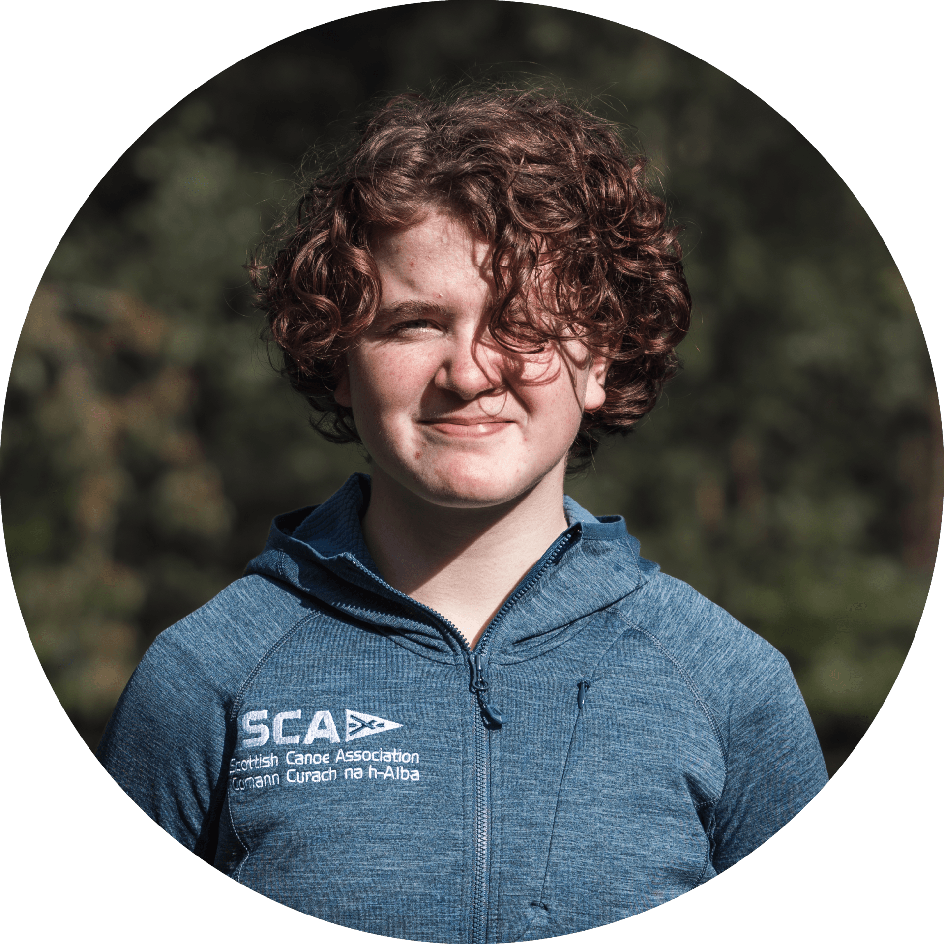 A young person with curly brown hair smiles in a blue zip-up jacket. Background is blurred greenery.