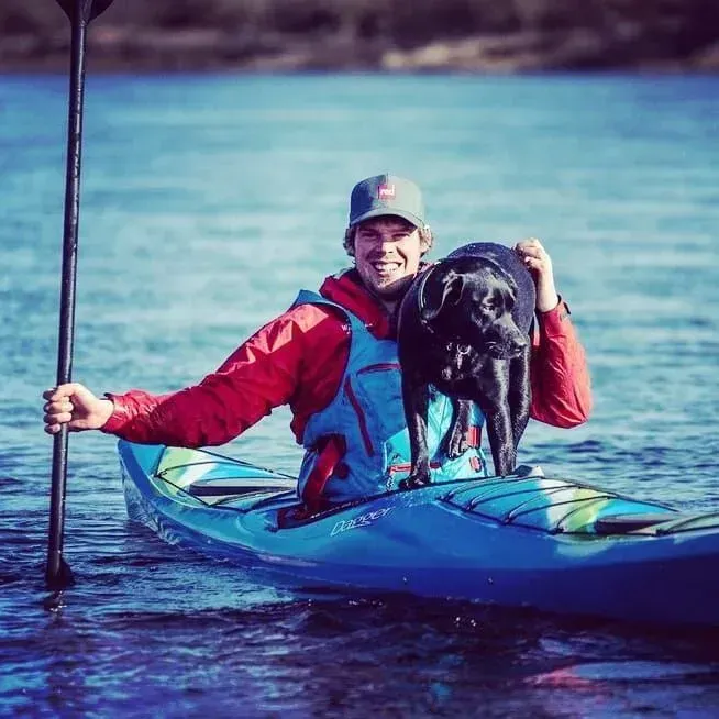 A man kayaking with a black dog on a lake; man is smiling and holding a paddle, dog is wet.