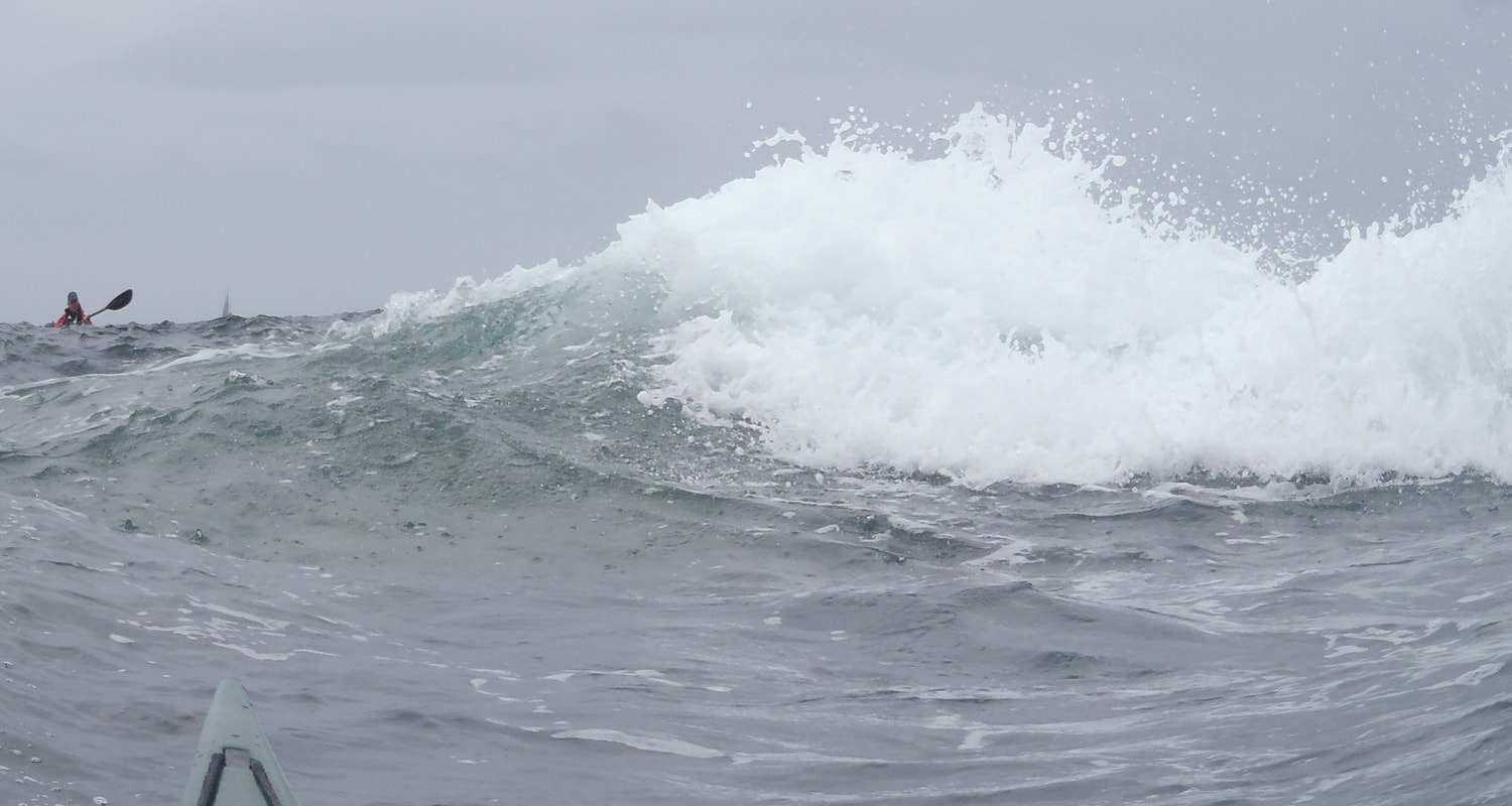 Large wave crashing in ocean with a kayaker in the background on a cloudy day.