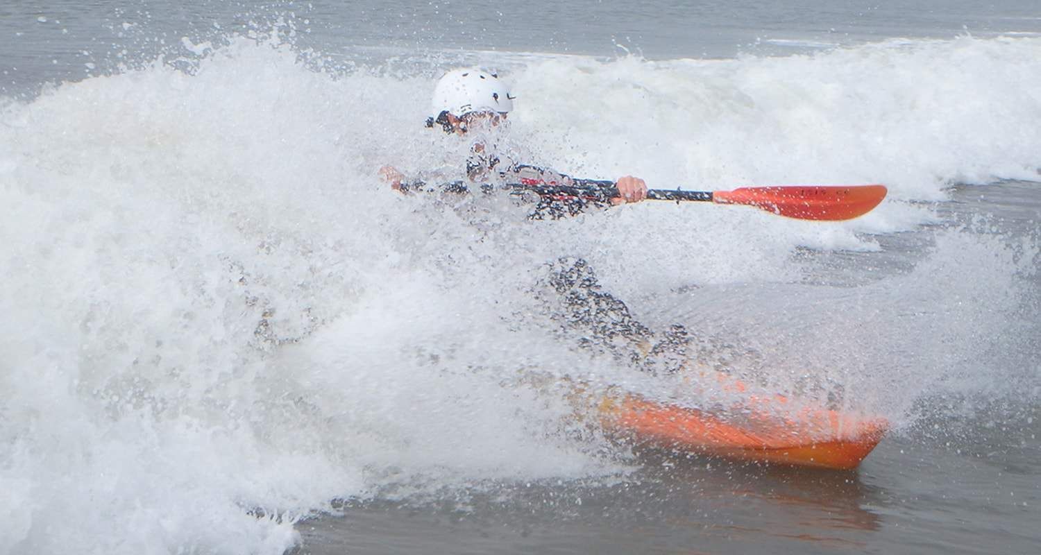 A kayaker navigates a wave, orange kayak amid white water, wearing a helmet and using a red paddle.
