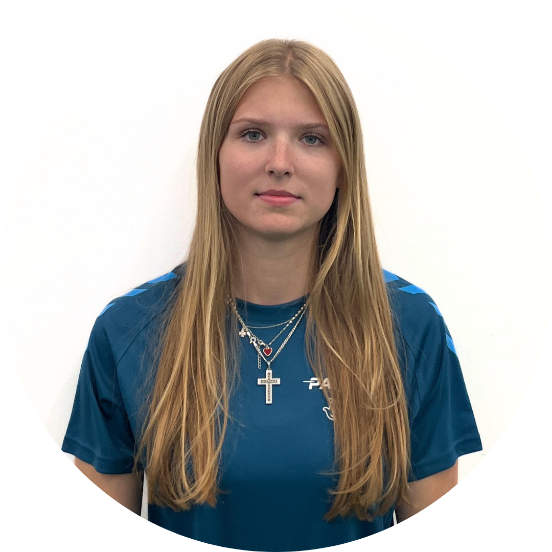 Blonde woman with long hair wearing a blue shirt and a cross necklace, standing in front of a white background.
