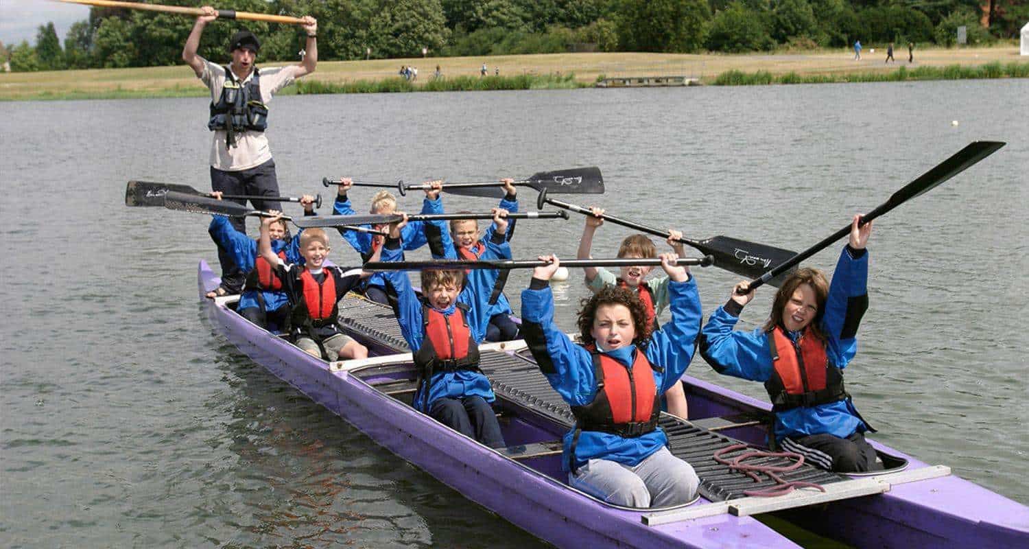 A group of children in blue jackets and life vests raise oars in a purple canoe on a lake.