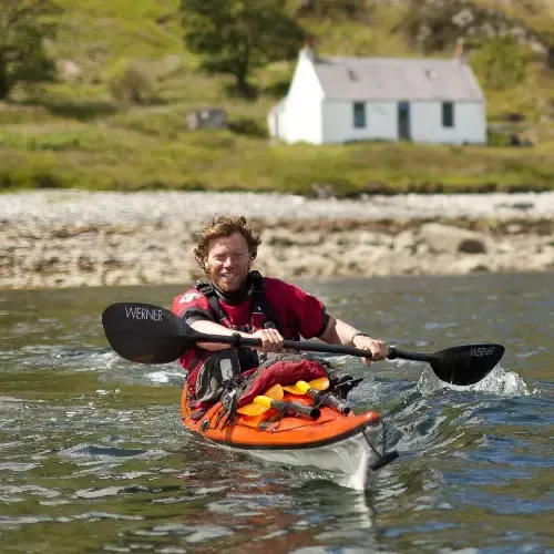 Man kayaking in orange kayak, red shirt, smiling. White cottage and green hillside in background.