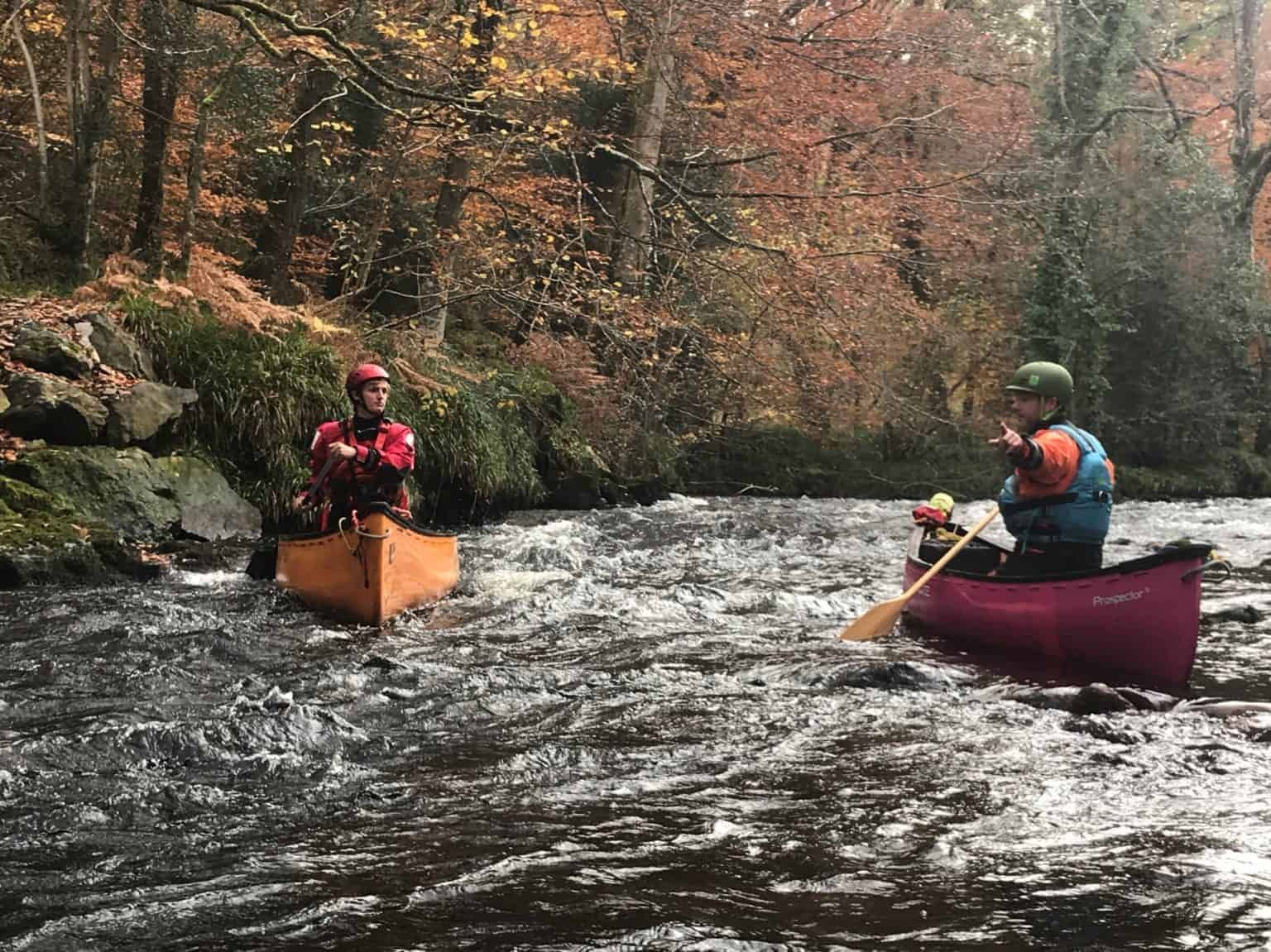 Two people canoe through a river with rapids, surrounded by autumn trees. One paddles, the other sits in the orange canoe.