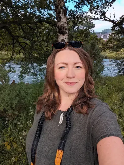 Woman with auburn hair smiles outdoors, wearing sunglasses and a necklace. A river and trees are in the background.