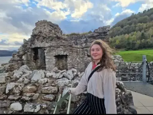 Woman smiling near ruins overlooking a lake, wearing a cream sweater and black pants. Cloudy sky in the background.
