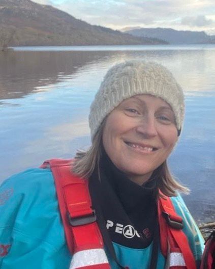 Woman in white beanie and life vest smiles, with a lake and mountains in the background.