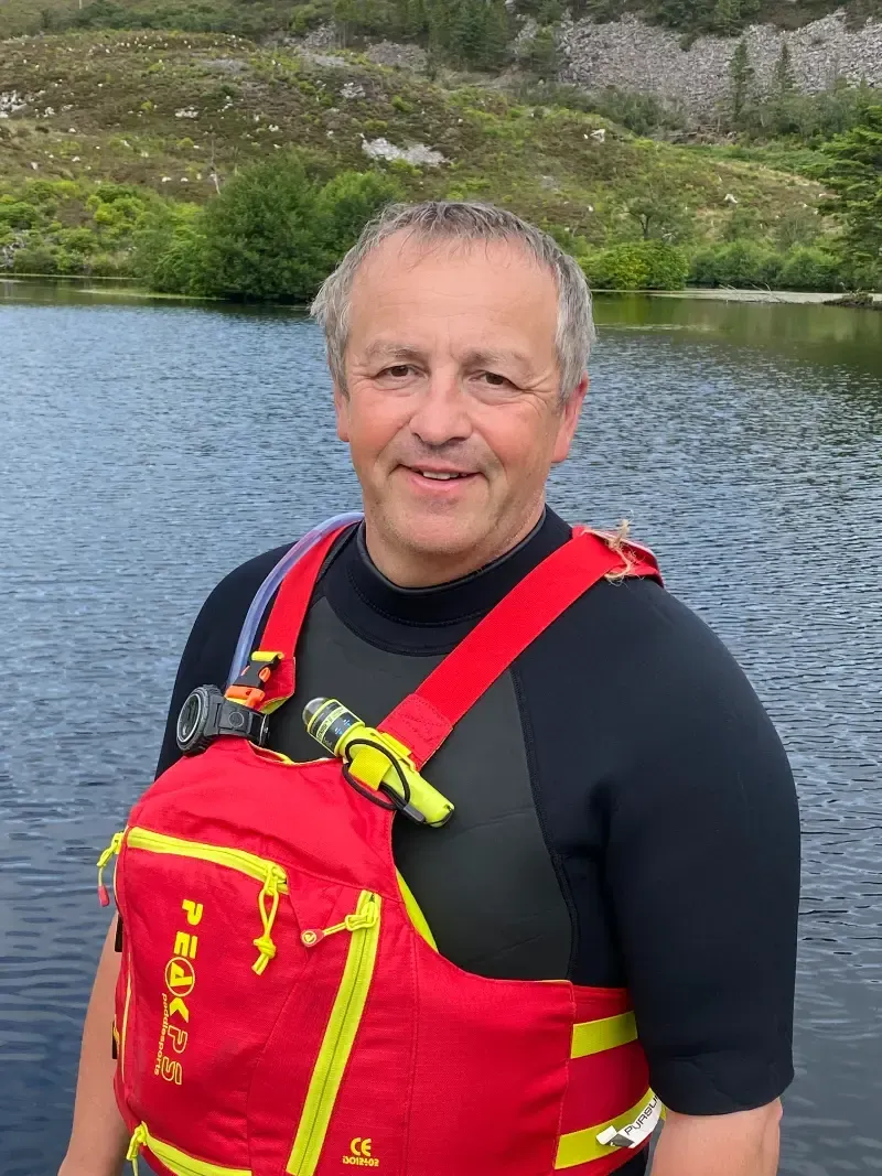 Man in red life vest and wetsuit smiles by a lake, with green hills in the background.