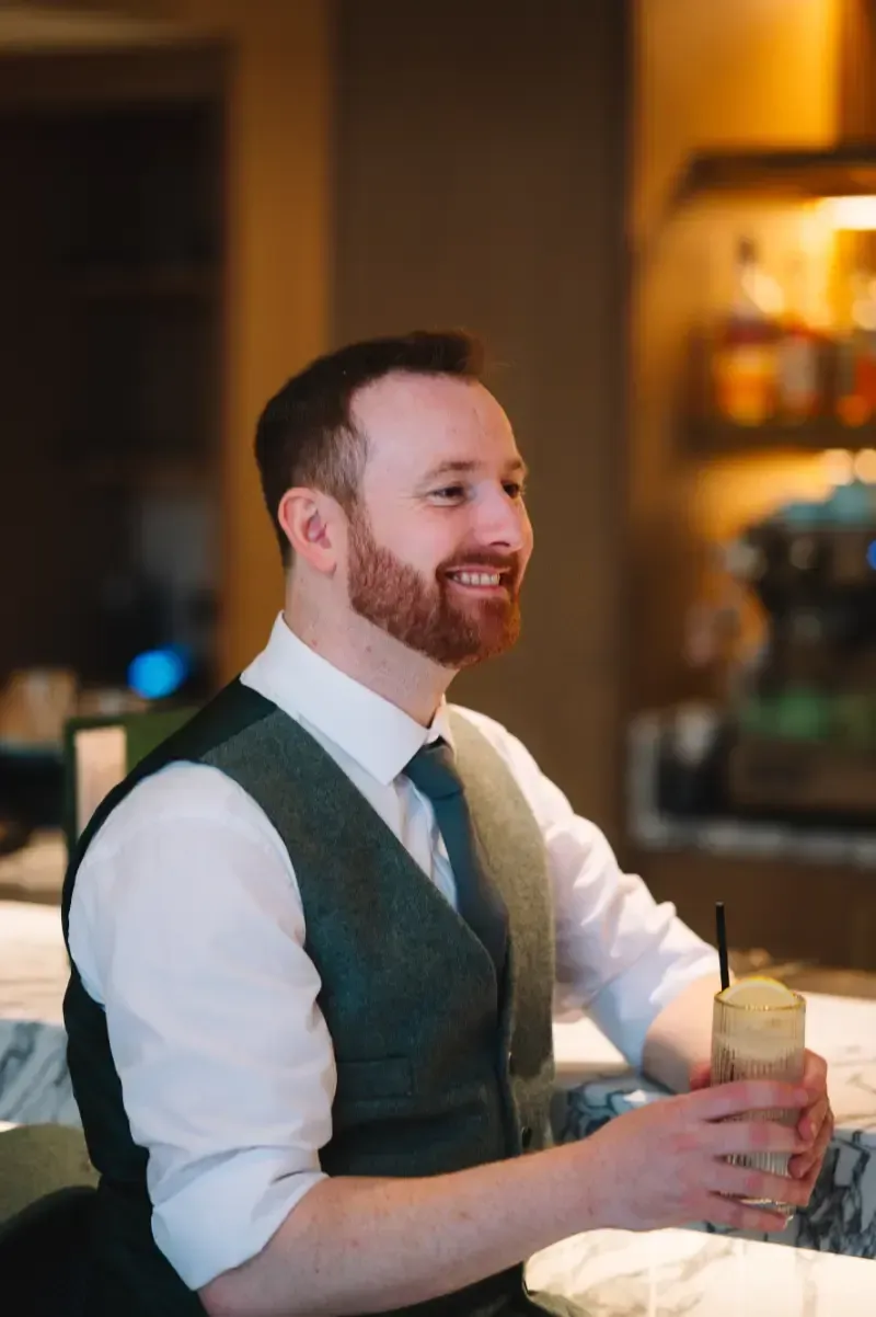A man with a red beard smiles, holding a drink at a bar. He wears a vest, tie, and white shirt.