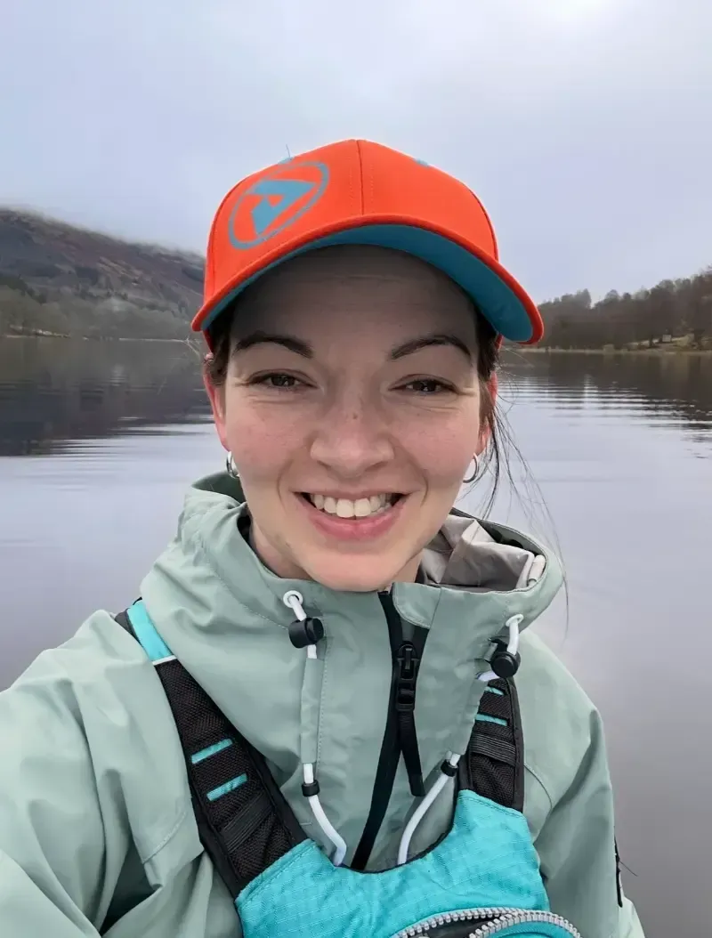 Woman in orange cap and light blue jacket smiles at the camera on a lake, foggy mountains in background.