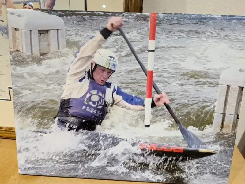 Canoe slalom racer navigates a gate in whitewater rapids. The athlete wears a helmet and paddles vigorously.