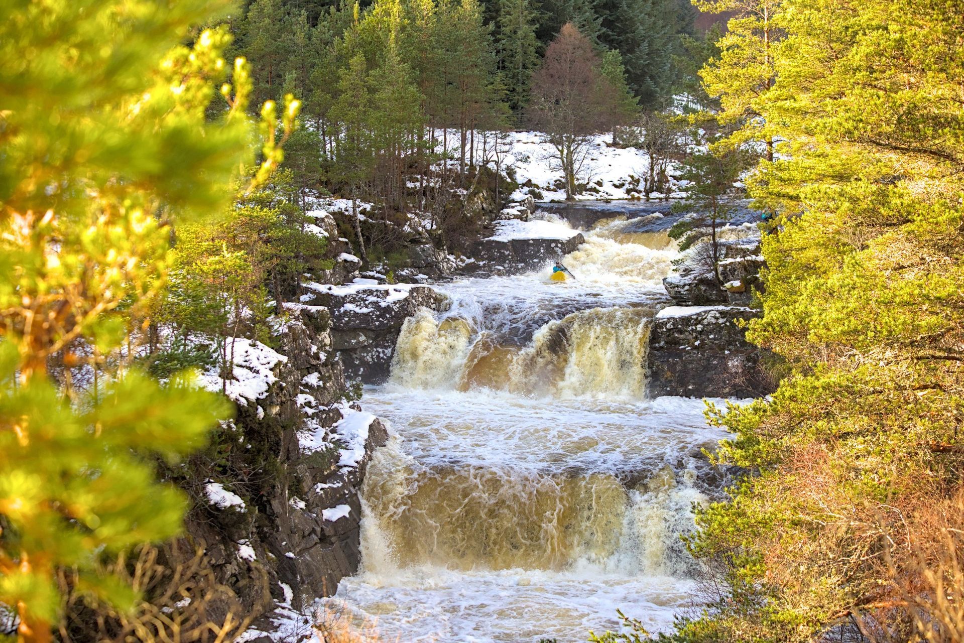 A kayaker wearing a blue helmet and colorful gear navigates whitewater rapids in a man-made channel, paddling forward with focused expression.