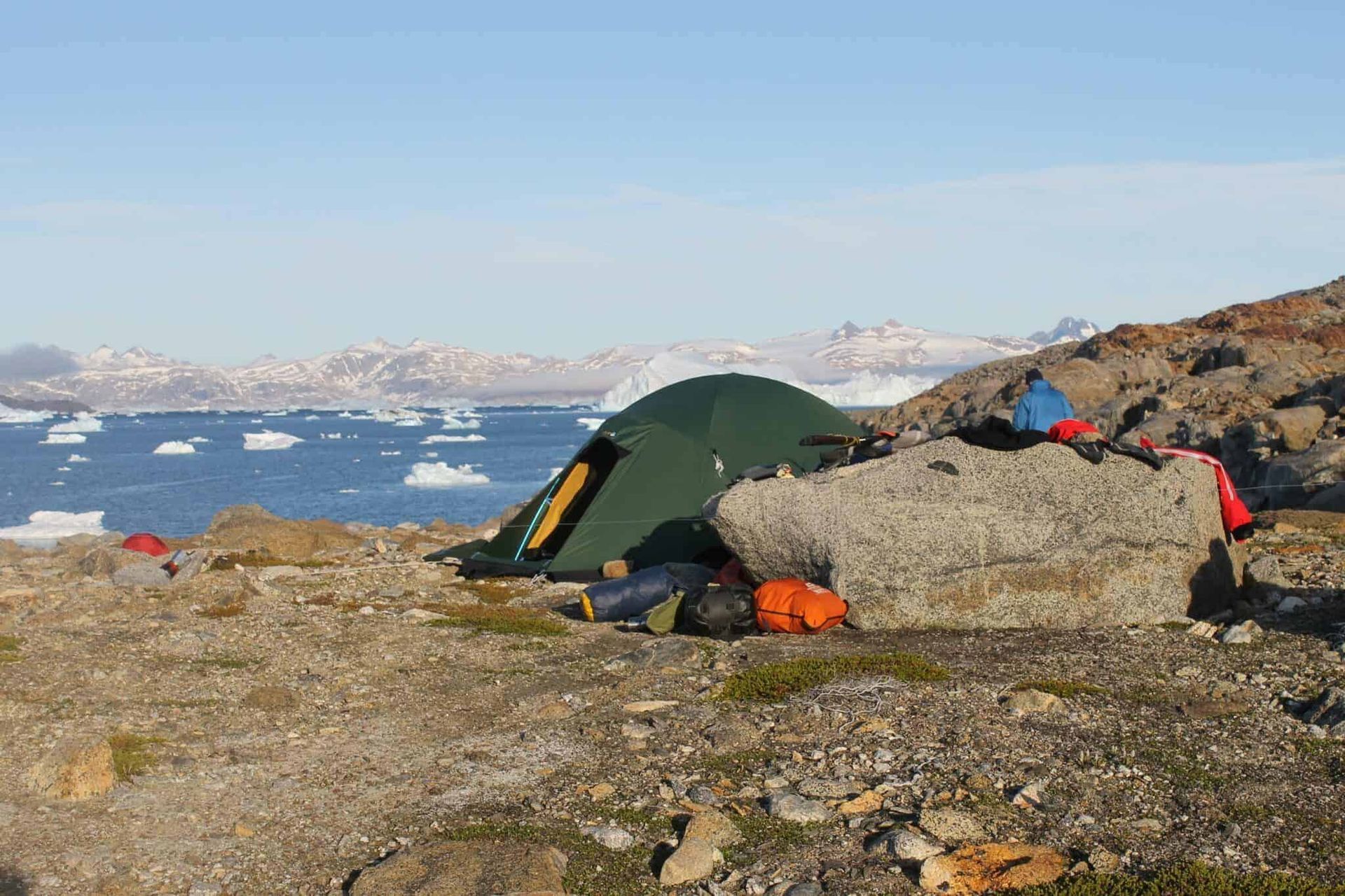 A green tent pitched on rocky terrain overlooking a sea filled with icebergs and mountains.