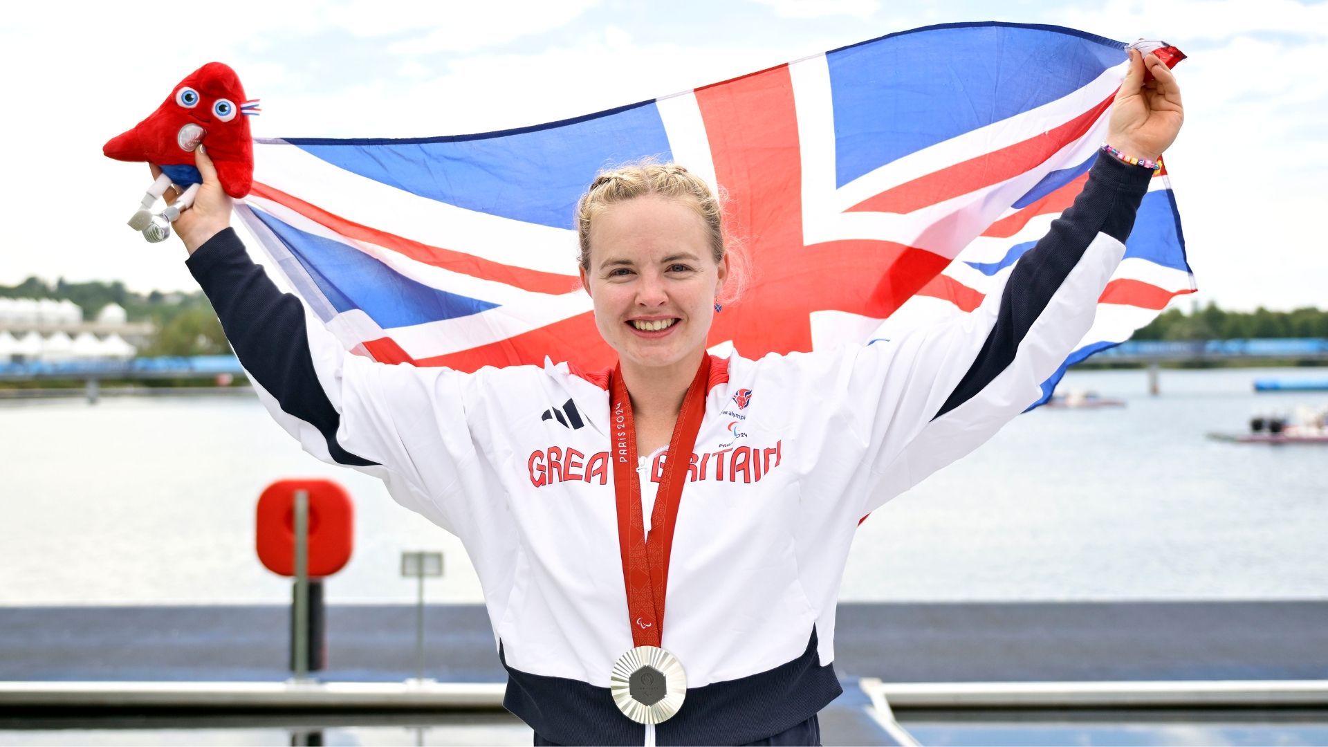 Female athlete in white and blue Great Britain uniform, holding up a silver medal and a Union Jack flag with a red plush toy.