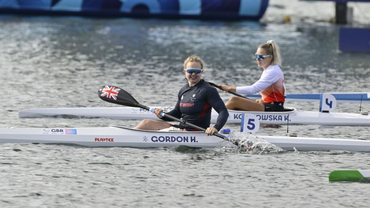 Two female kayakers racing on a lake. The lead athlete, wearing a Great Britain top, paddles forward. The other, in an orange shirt, follows behind.