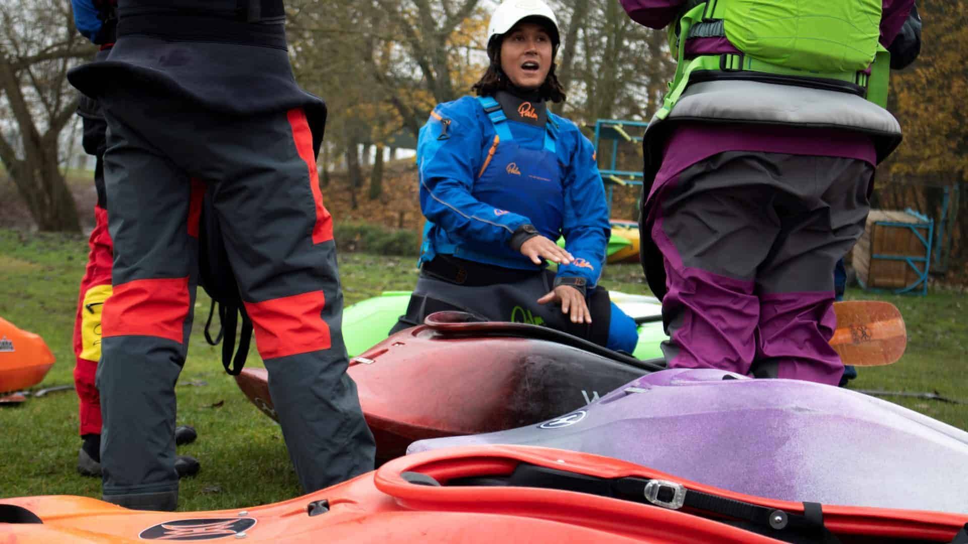 A woman in blue giving instructions near kayaks. She wears a white helmet and gestures with her hands while others listen.
