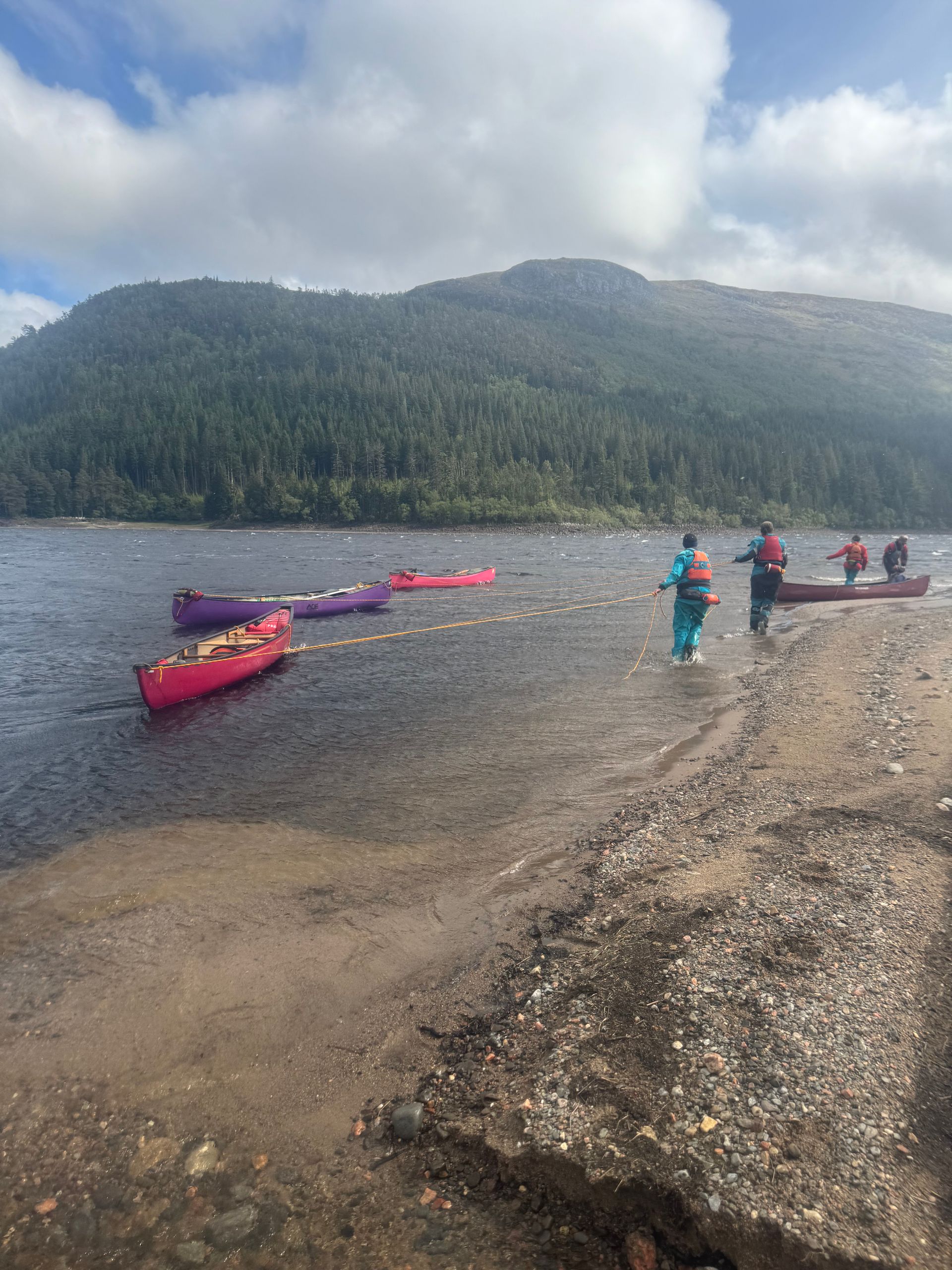 People prepare to canoe on a lake with a forested mountain backdrop. Colorful canoes are on the shore.