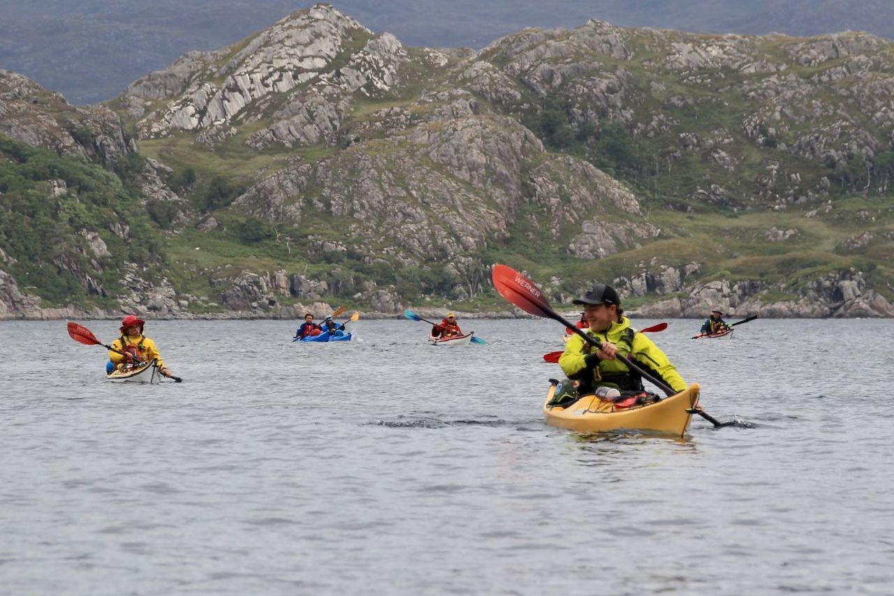 Kayakers on a body of water near a mountainous landscape. Several paddlers use red paddles in yellow and blue kayaks on a cloudy day.