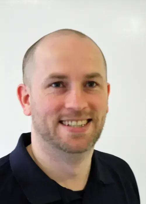 Bald man with fair skin smiles, wearing a dark blue collared shirt, against a white background.