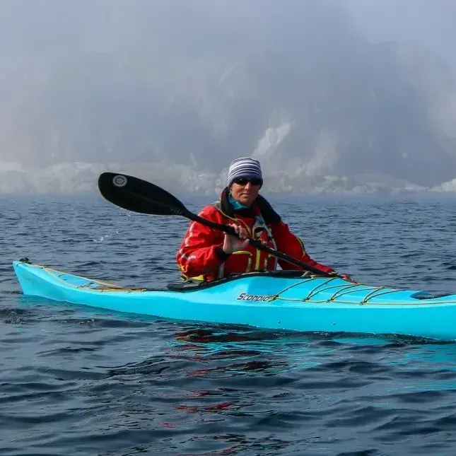 Person kayaking in a blue kayak on calm water; red jacket, foggy background.