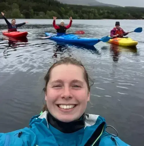 Woman taking a selfie with three people kayaking in a lake. They are smiling and waving in front of a forested shoreline.