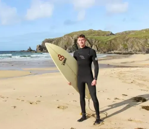 Man in black wetsuit holding a surfboard on a sandy beach, with a rocky coastline in the background under a partly cloudy sky.