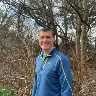 Man in blue athletic jacket smiles outdoors, standing near bare trees and bushes.