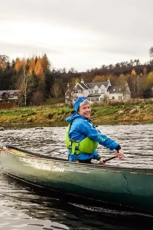 Person in a canoe wearing a blue jacket and life vest, paddling on a river with buildings and trees in the background.