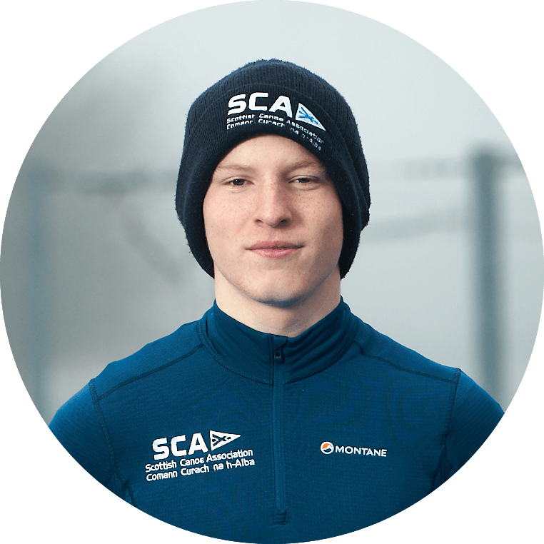 A young man wearing a beanie and blue athletic shirt with the Scottish Canoe Association logo smiles.