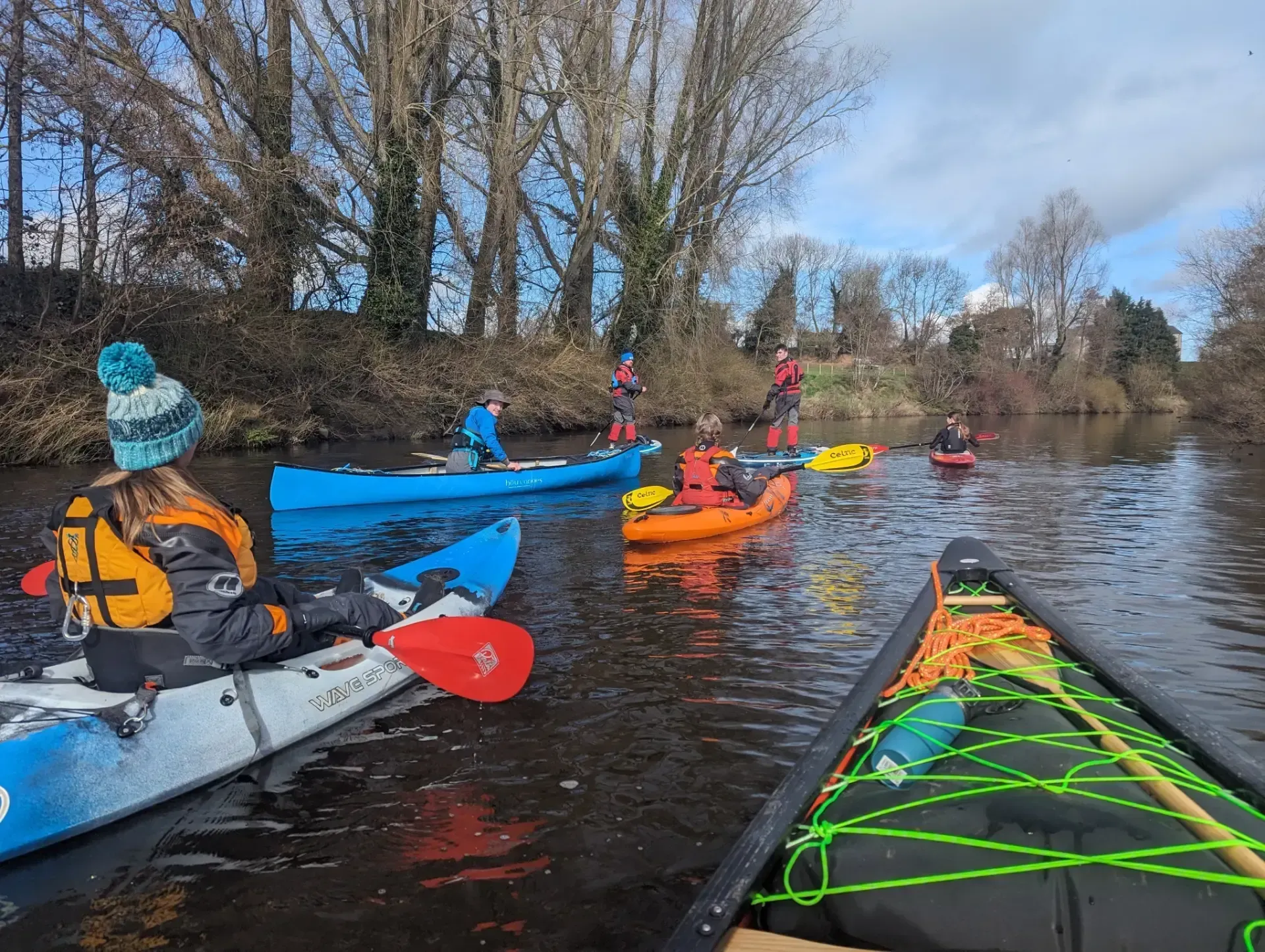 People kayaking on a river under a cloudy sky. Several colorful kayaks are on the water, with individuals wearing life vests. Bare trees line the riverbank.