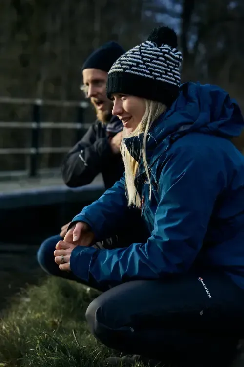 Woman in blue jacket and patterned hat squats outdoors, looking to the side, man in black hat behind her, mouth open.