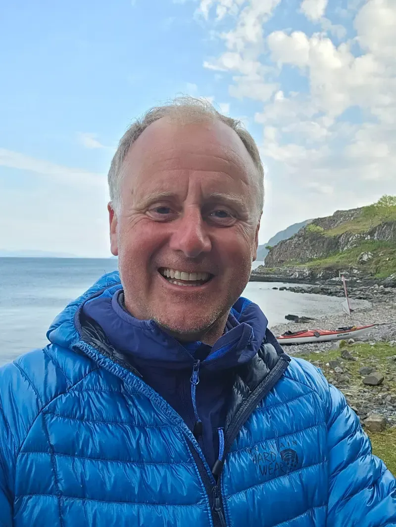 Smiling man in a blue jacket outdoors near a rocky coastline with kayaks, bright blue sky.