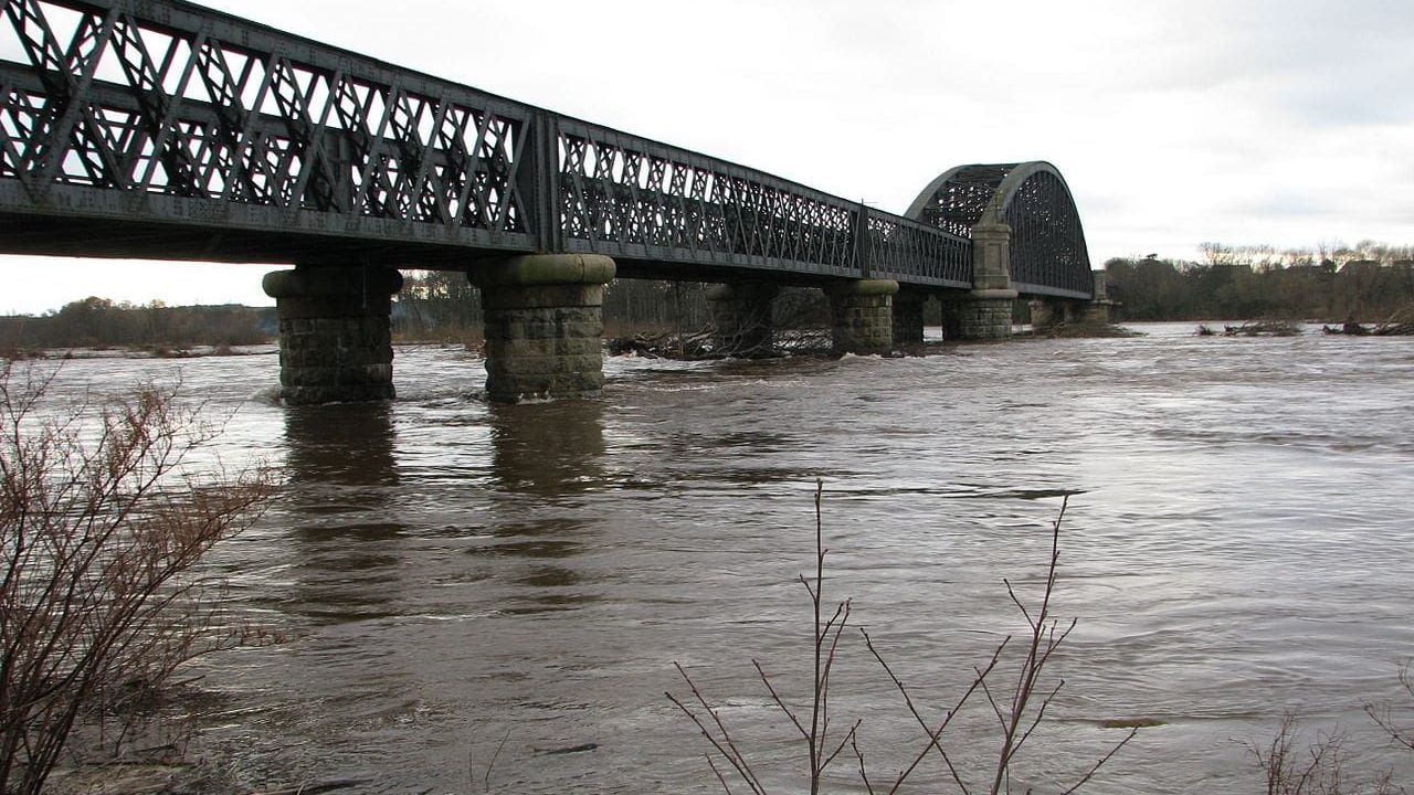 An old metal bridge arches over a wide, muddy river. The overcast sky reflects in the water.
