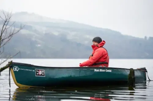 Person in red jacket sits in a green canoe on a lake, with a misty mountain in the background.