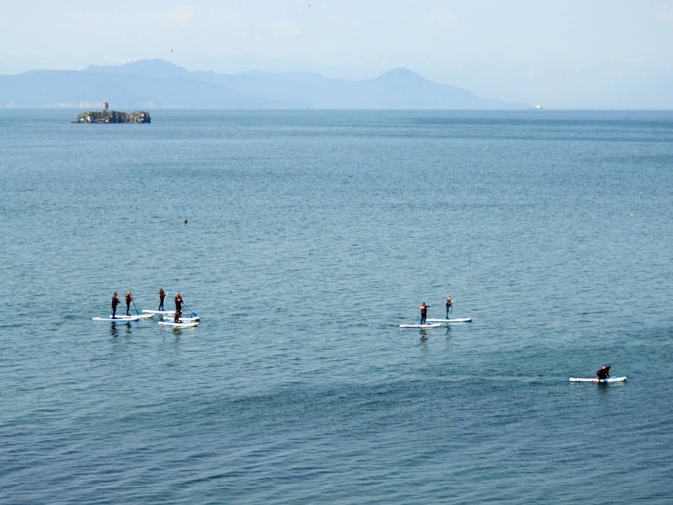 People paddleboard on a blue ocean. In the distance, a small island and a mountain range are visible under a hazy sky.