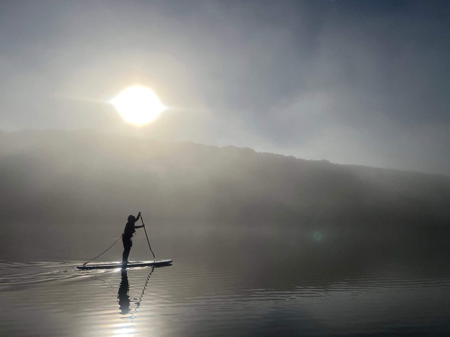 A person paddleboarding on calm water under a bright sun, with mountains in the background obscured by mist.