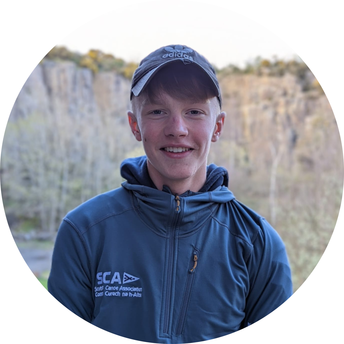 A young man with fair skin wearing a cap and blue jacket smiles in front of a rocky cliff.